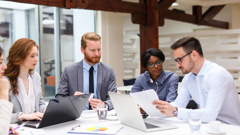 A group of people in a business meeting is working together at a table with laptops and documents.