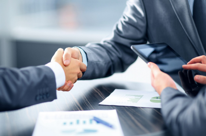 Two people in business suits shake hands over a table with documents and a tablet.