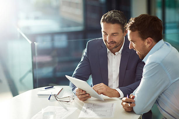 Two men in business attire discussing documents and a tablet in an office setting.