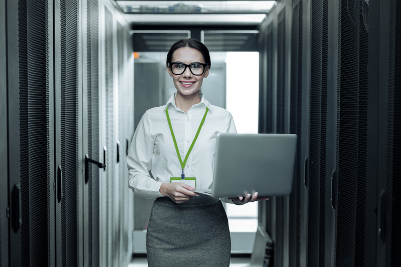 A person wearing glasses and a lanyard works on a laptop while standing in a server room.